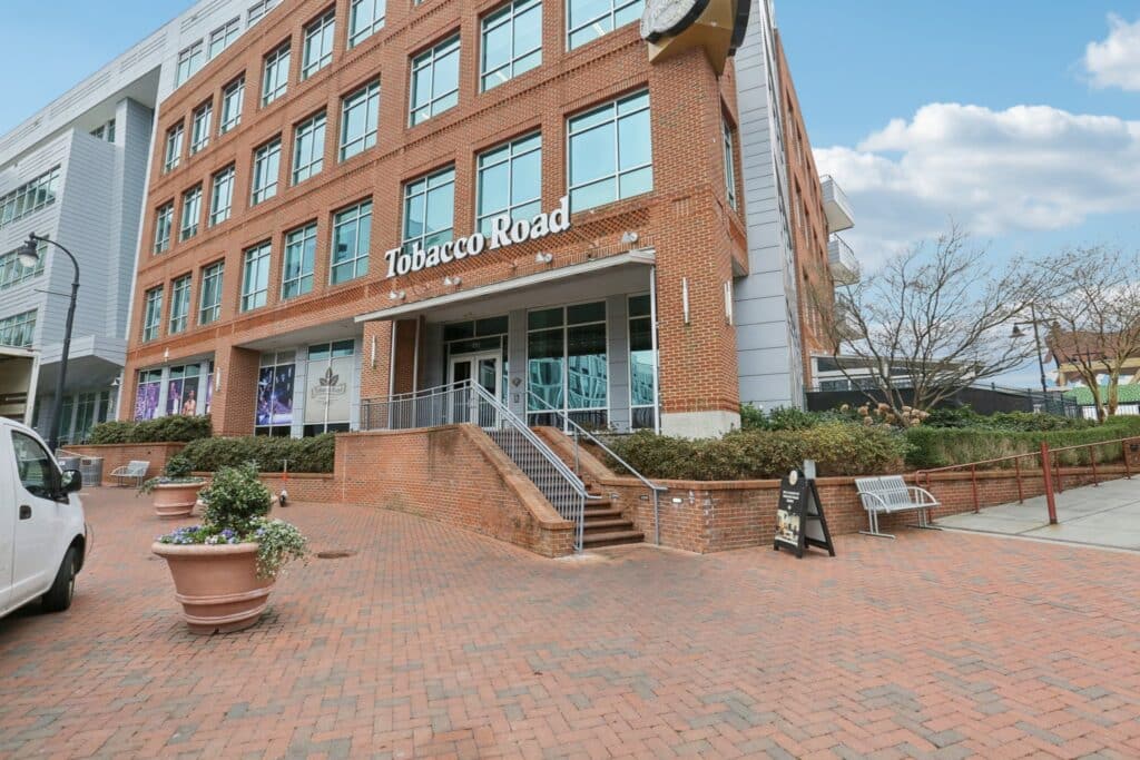 Brick building labeled "Tobacco Road" with steps leading to the entrance, surrounded by large plant pots, on a clear day.