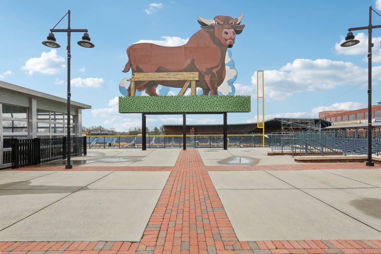 Outdoor stadium area with large bull billboard and empty seating. Brick pathway leads to stands. Stadium lights and blue sky in the background.