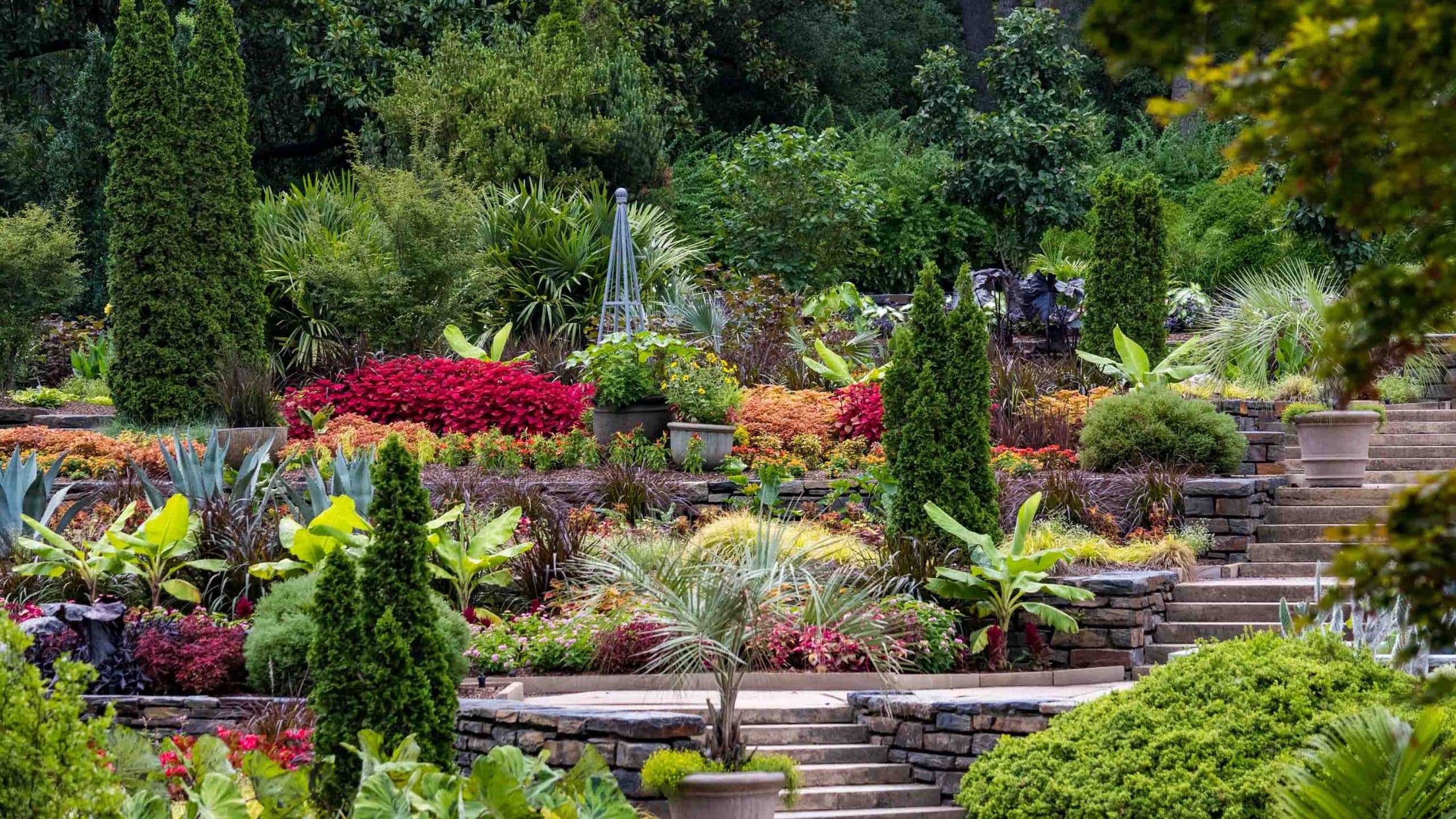 A tiered garden with stone steps features diverse greenery, colorful flowers, and tall shrubs, surrounded by dense trees in the background.